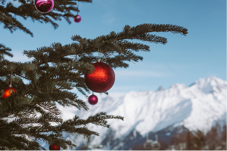 Christmas tree branches decorated with red and pink baubles, with snow-covered Alpine mountains in the background.