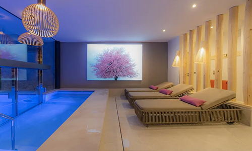 Modern Alpine hotel spa featuring an illuminated indoor swimming pool, comfortable loungers and warm wooden interior details.